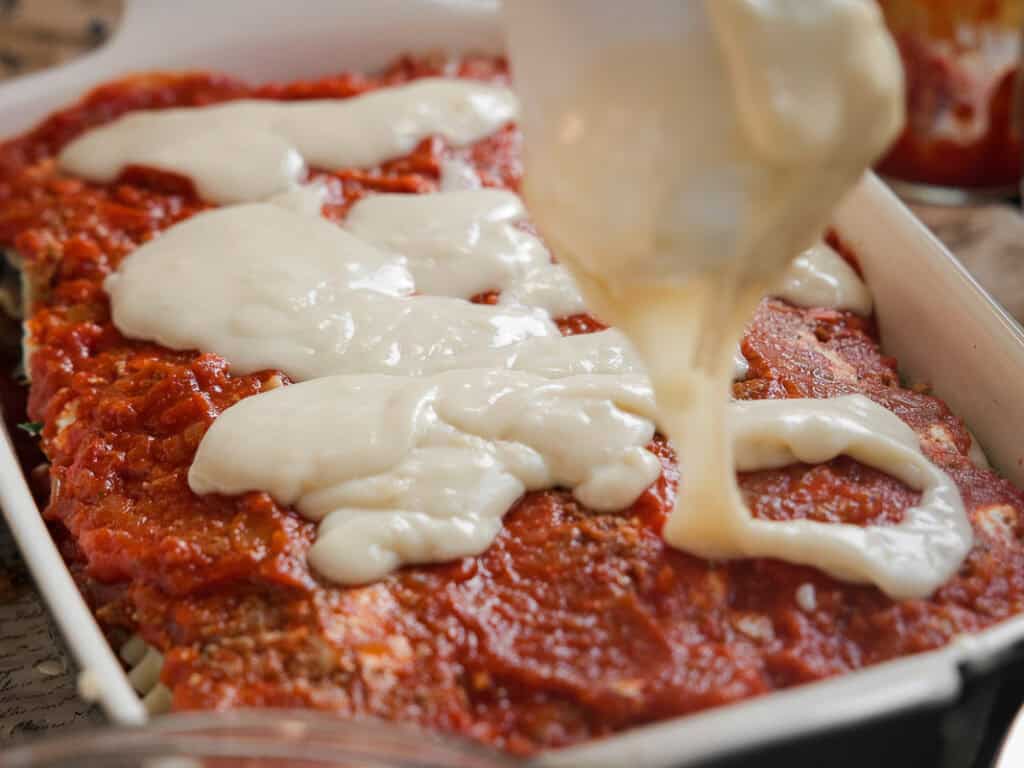 Close-up of a baking dish filled with layers of vegan lasagna. A hand is pouring creamy white sauce over a layer of chunky red tomato sauce, creating a rich and layered effect. The image captures the preparation process of the dish.